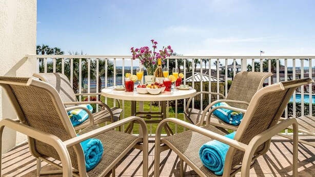 A balcony with wicker chairs, a table with drinks and food, and a view of the pool and ocean. - 🌴Welcome to The Islander Unit #203 Your Perfect Destin Beach Escape🌅 (Destin)