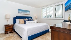 A bedroom with a bed, nightstands, a dresser, a window with blinds, and a painting of a beach scene. - 🌴Welcome to The Islander Unit #203 Your Perfect Destin Beach Escape🌅 (Destin)