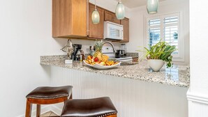 A kitchen with wooden cabinets, a granite countertop, a microwave, a sink, a fruit bowl, two bar stools, and pendant lighting. - 🌴Welcome to The Islander Unit #203 Your Perfect Destin Beach Escape🌅 (Destin)