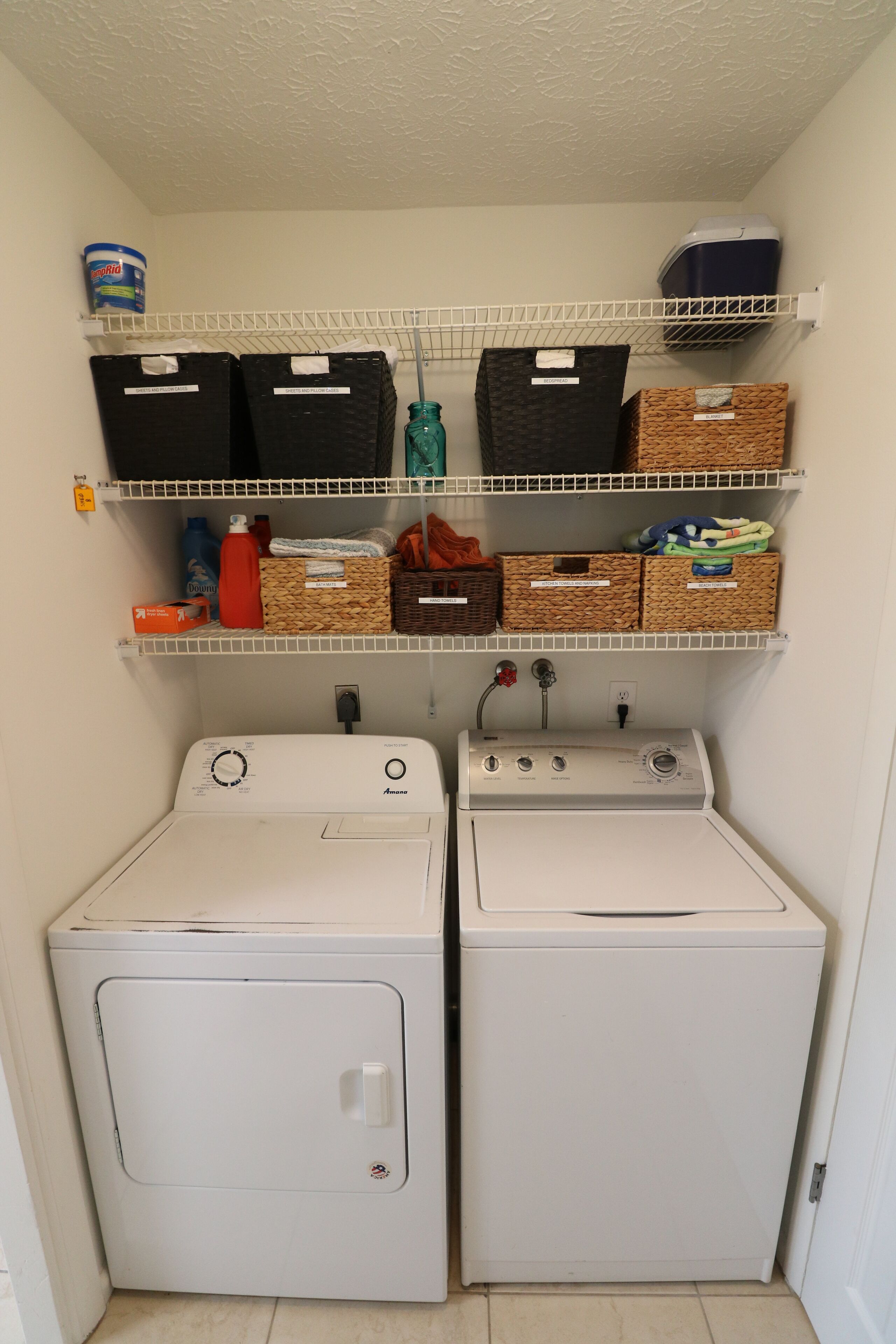 A laundry room with two front-loading washing machines and three shelves above containing baskets and storage boxes.