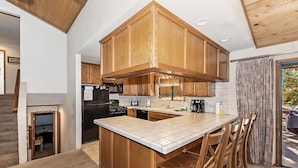 A kitchen with wooden cabinets, a central island, and a dining area with chairs. - Fox Canyon by Destination Big Bear : Hot Tub! BBQ! Air Hockey! Internet! Deck! Great Neighborhood! Fireplace! (Big Bear Lake)