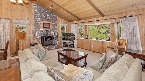 A cozy cabin interior with a stone fireplace, wooden walls, a ceiling fan, and a large window offering a view of trees. - Fox Canyon by Destination Big Bear : Hot Tub! BBQ! Air Hockey! Internet! Deck! Great Neighborhood! Fireplace! (Big Bear Lake)