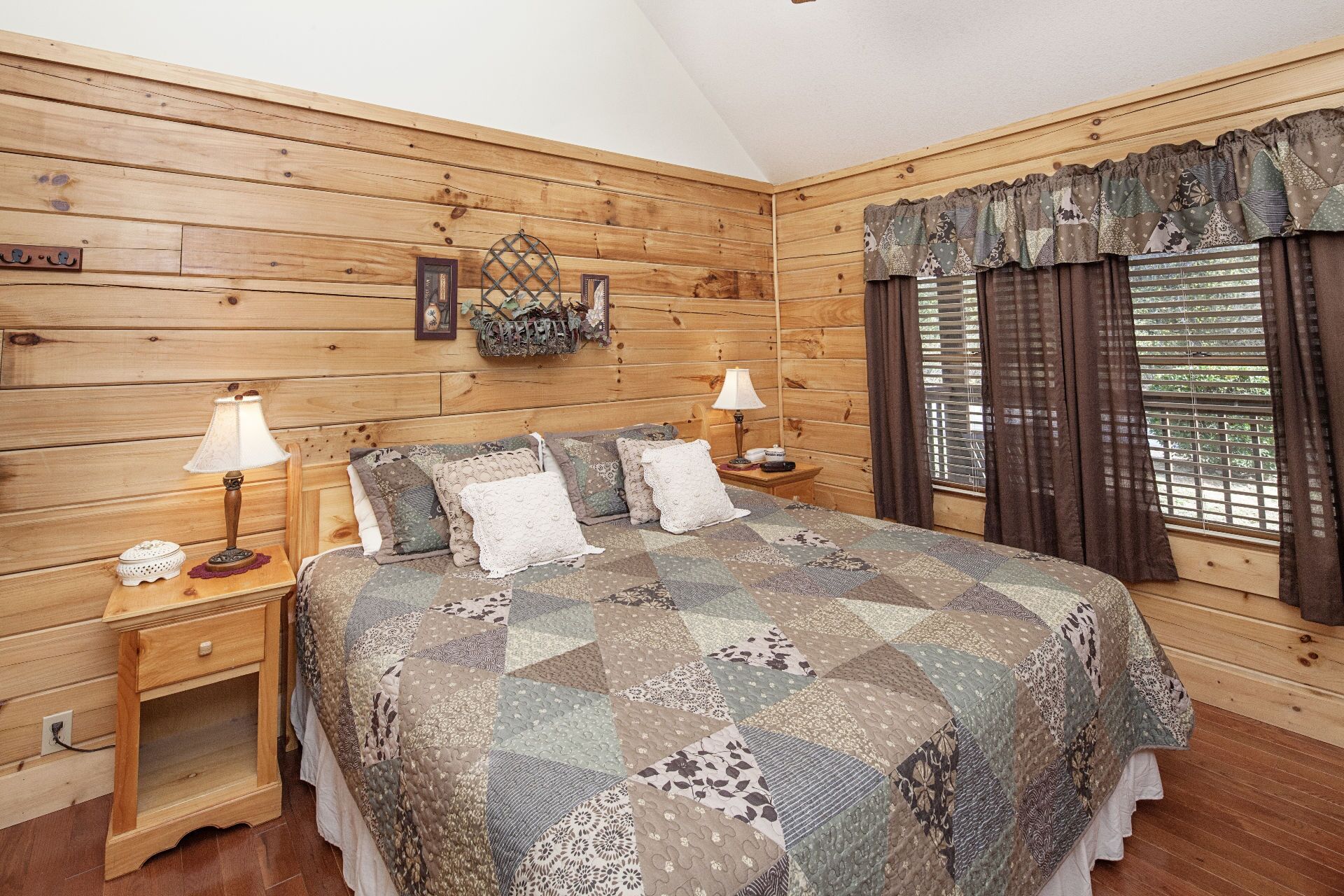 A cozy bedroom with a wooden log wall, a large bed with a patchwork quilt, two bedside tables with lamps, and a window with curtains.