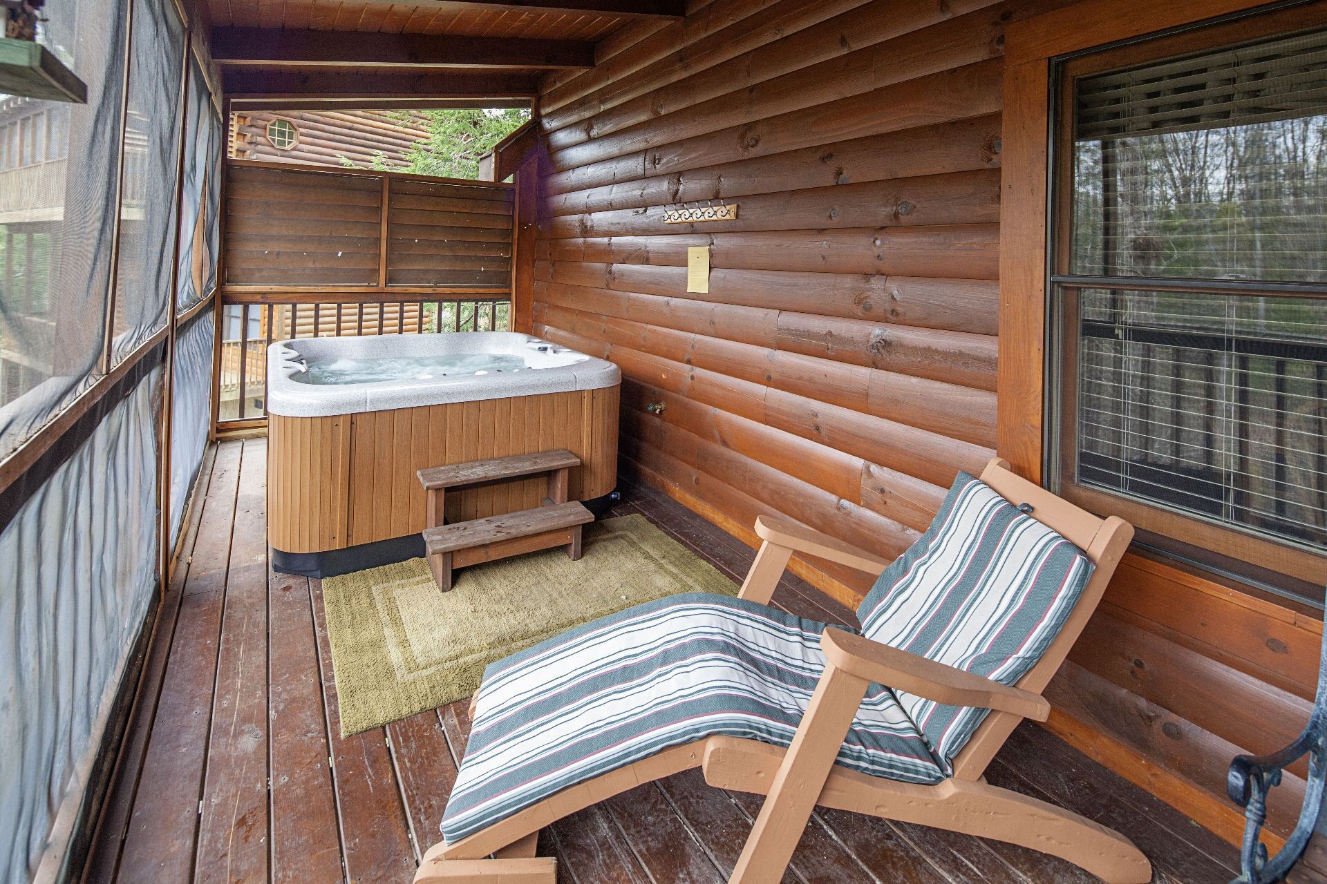 A wooden cabin with a hot tub, a wooden deck, and a window with blinds.