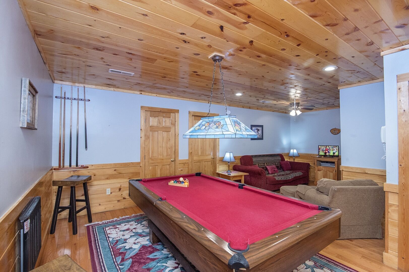 A pool room with a pool table, wooden walls, a red carpet, and a cozy seating area with a sofa and armchairs.