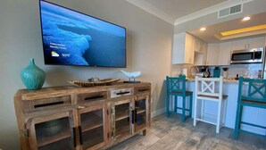 Een moderne keuken met witte kasten, teal stoelen en een houten kastje met een televisie waarop het weer wordt getoond.