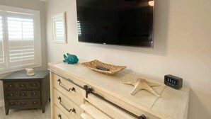 Een kamer met een wandtelevisie, een dressoir met lades, een tafeltje met een afstandsbediening en een decoratie, en een zeester decoratie op het dressoir.