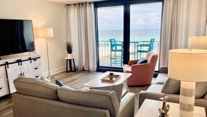 Habitación de hotel moderna con un televisor grande, asientos cómodos, una mesita con bandeja y vistas al océano a través de las puertas del balcón.
