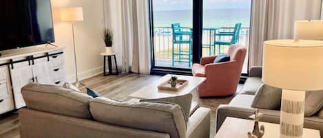 A modern hotel room with a large TV, comfortable seating, a small table with a tray, and a view of the ocean through the balcony doors.