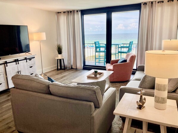 A modern hotel room with a large TV, comfortable seating, a small table with a tray, and a view of the ocean through the balcony doors.