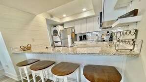 Una cocina moderna con armarios blancos, lavadora integrada y encimera de mármol. Hay tres taburetes de madera con barras de metal.