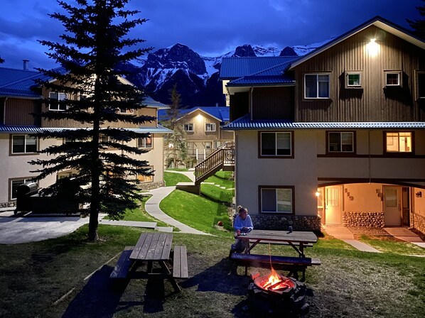 Un chalet doté d’un foyer extérieur et d’une table de pique-nique dans la cour, entouré de bâtiments et de montagnes.