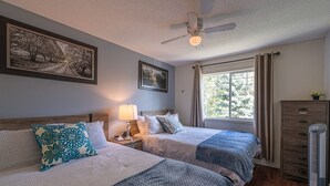 A bedroom with two beds, a ceiling fan, a nightstand with a lamp, a dresser, and a window with curtains. - Gorgeous view 2 Storey Townhouse 2 BR/1.5 Bath, A/C, Hot Tub, Fire Pit  (Harvie Heights)