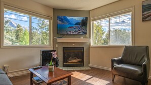 A living room with a fireplace, a television mounted above it, and a large window offering a view of mountains and trees. - Gorgeous view 2 Storey Townhouse 2 BR/1.5 Bath, A/C, Hot Tub, Fire Pit  (Harvie Heights)