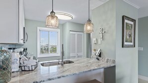 A kitchen with a marble countertop, two pendant lights, a sink, and a window with blinds. - Emerald Coast Vibes & Sunset Views – Islander 517 (Destin)