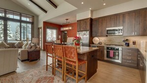 A modern kitchen with dark wood cabinets, stainless steel appliances, and a large island. There is a dining area with a sofa and chairs, and a large window providing a view of snow-covered trees. - Gondola Ski-in/Ski-out By Tahoe Getaways - Luxury 4 BR, Private Hot Tub, Access to Pool (Truckee)