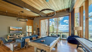 A modern living room with a pool table, comfortable seating, and large windows offering a view of the outdoors. - Gondola Ski-in/Ski-out By Tahoe Getaways - Luxury 4 BR, Private Hot Tub, Access to Pool (Truckee)