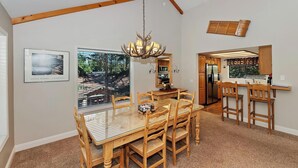 A dining area with a glass-top table and wooden chairs, a chandelier, and a kitchen visible through a window.