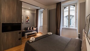 Un dormitorio moderno con una cama grande, televisor, zona de estar con sofá y mesa de centro, y ventana con cortinas.