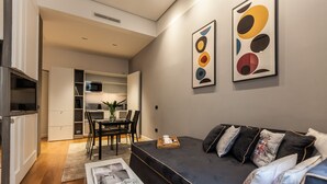 Una sala moderna con un sofá oscuro, una mesa de centro y un comedor con sillas y una mesa. Se observa una zona de cocina con armarios y un microondas.