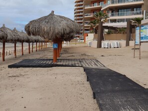 Uma área de praia com guarda-sóis de palha, um sinal indicando acessibilidade para cadeiras de rodas e um edifício de vários andares ao fundo.