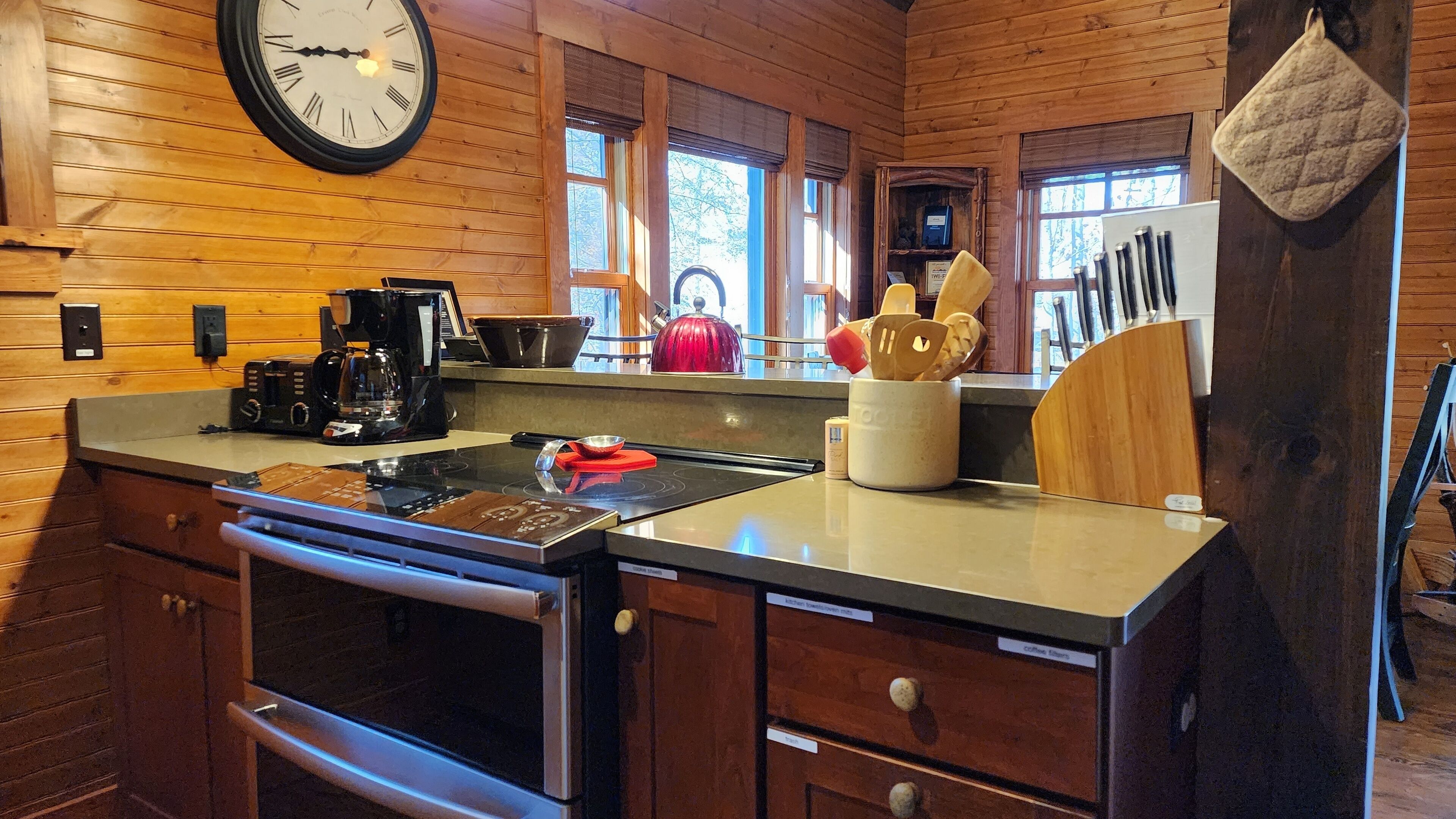 A kitchen with wooden walls, a clock, a stove, a coffee maker, and various kitchen utensils.