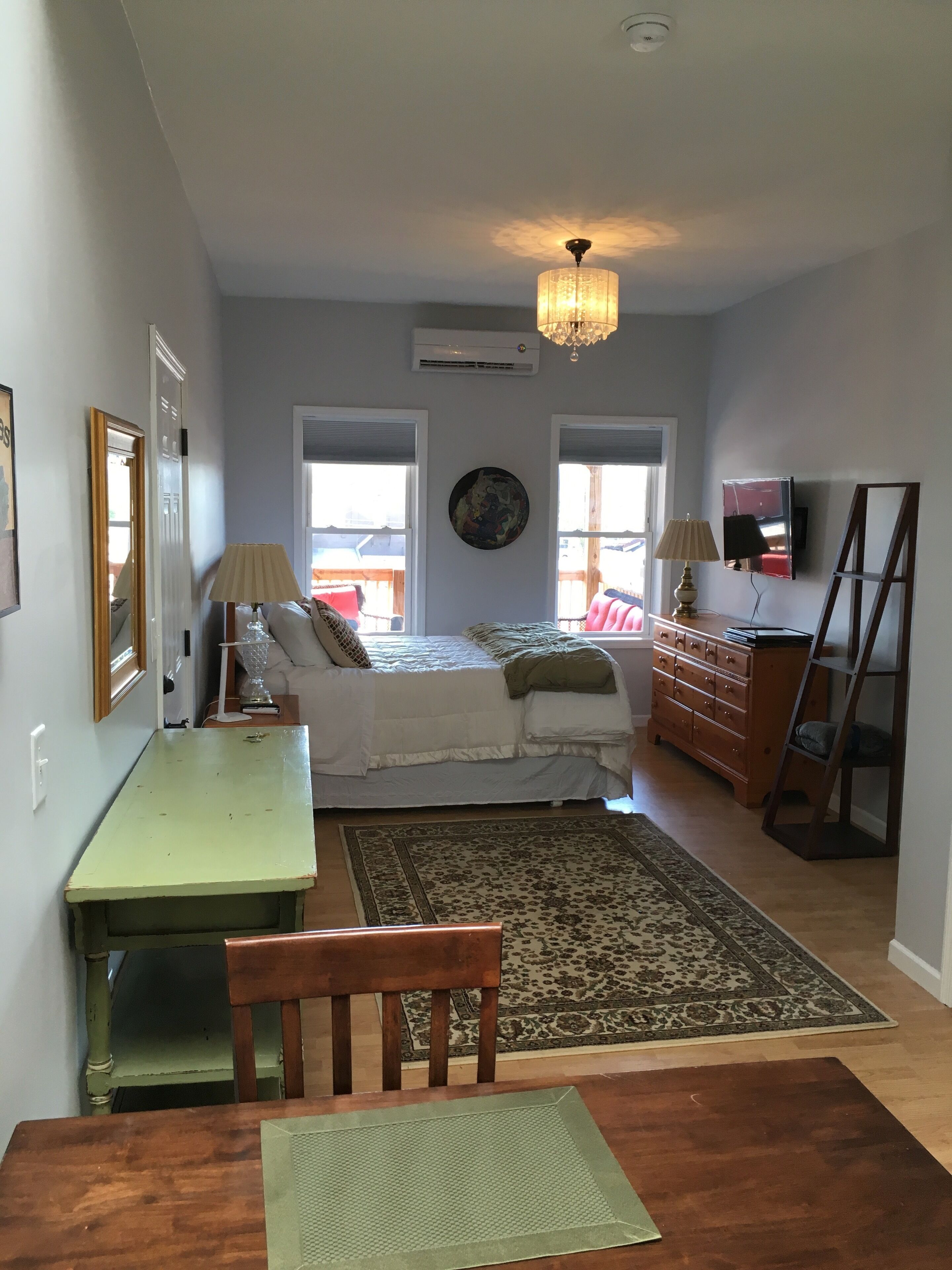 A bedroom with a bed, dresser, and a desk with a chair. There is a window, a ceiling fan, and a TV on the wall.