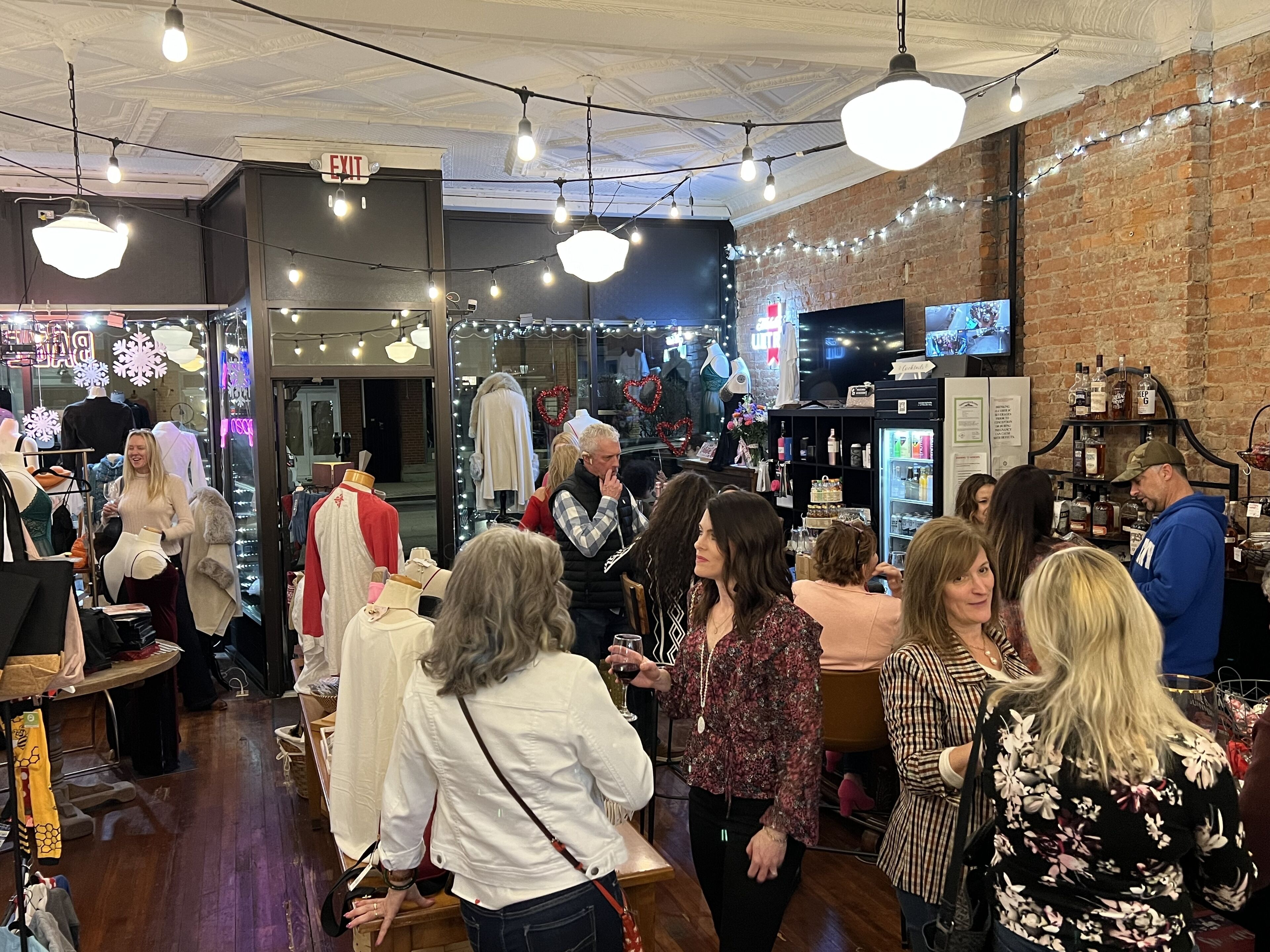 A crowded indoor space with mannequins displaying clothing, a bar area with bottles, and a brick wall.
