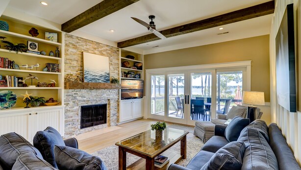 A spacious living room with a stone fireplace, a large TV, and a view of the outdoors through sliding glass doors. - 9 Lands End Rd-Pretty view, Pool Onsite, Easy Walk to water & S. Beach Marina (Hilton Head Island)