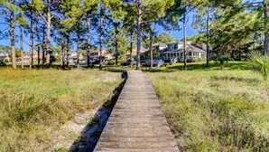 A wooden boardwalk leading to a house with a covered porch, surrounded by pine trees and a grassy area. - 9 Lands End Rd-Pretty view, Pool Onsite, Easy Walk to water & S. Beach Marina (Hilton Head Island)