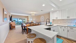 A modern kitchen with white cabinetry, a central island, and a dining area with wicker chairs. - 442 Captains Walk | Direct Oceanfront Views (Hilton Head Island)