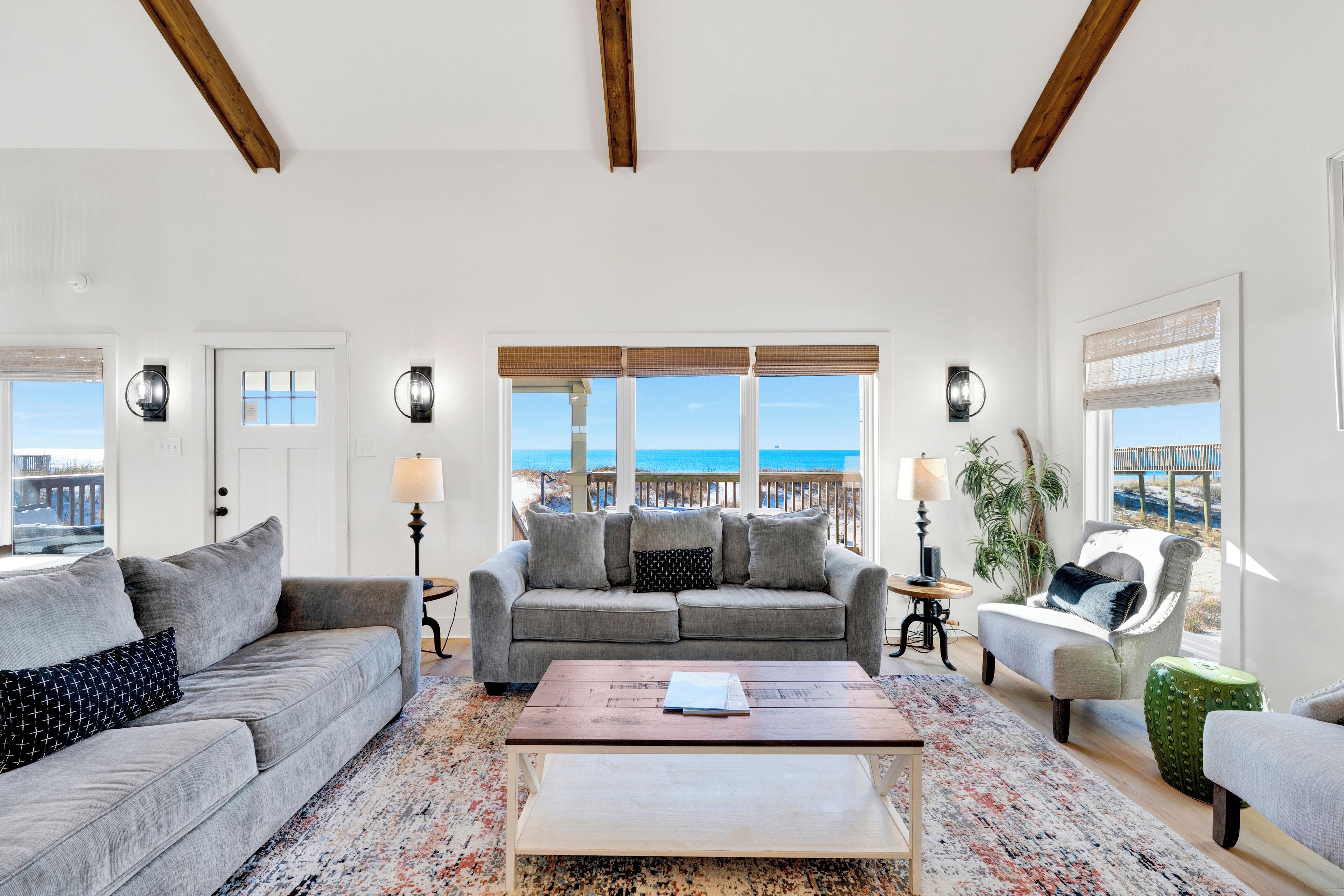 A spacious living room with a large window offering a view of the beach, a grey sofa, a coffee table, and a chair with a view.