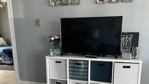 A modern living room with a flat-screen TV, a white entertainment unit with drawers, and a decorative wall piece above the TV. - Waterfront Condo Within Steps To Wells Beach!  (Wells)