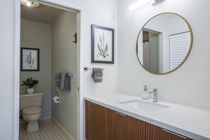 A modern bathroom with a toilet, sink, and mirror. There are framed botanical prints on the wall and a towel rack with towels. - Beautiful Ocean Views From Maui Kamaole I-118   (Kihei)