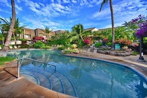 A swimming pool with a clear blue water feature, surrounded by palm trees and tropical plants. - Beautiful Ocean Views From Maui Kamaole I-118   (Kihei)
