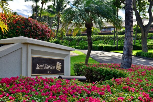 A sign with the name "Mai Kamole" and a number "2777" in a landscaped area with pink flowers and palm trees. - Beautiful Ocean Views From Maui Kamaole I-118   (Kihei)