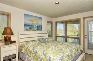 A bedroom with a bed, a nightstand, a lamp, a painting, and windows.