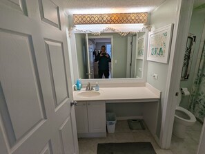 Un baño con un tocador blanco, un espejo grande, un perchero, un cubo de basura y un inodoro.