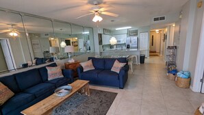 A modern living room with a dark blue sofa, a wooden coffee table, and a patterned rug. A kitchen with white cabinets and a dining area with a table and chairs is visible in the background. - Breathtaking views directly facing the Gulf!  3bd/3bath/6 (Fort Walton Beach)