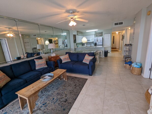 A modern living room with a dark blue sofa, a wooden coffee table, and a patterned rug. A kitchen with white cabinets and a dining area with a table and chairs is visible in the background. - Breathtaking views directly facing the Gulf!  3bd/3bath/6 (Fort Walton Beach)