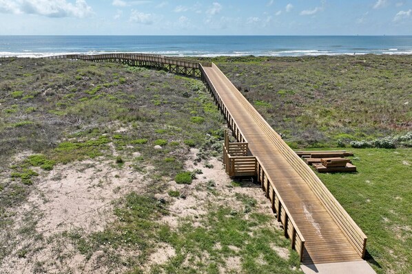 Una pasarela de madera que se extiende sobre una playa arenosa con vegetación escasa, y que lleva a una zona cubierta de césped con bancos.