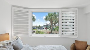 Ein Schlafzimmer mit großem Fenster, einem Bett mit grauer Bettwäsche, einem braunen Sessel und Blick auf einen Parkplatz und Palmen.