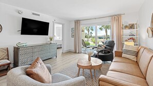 Ein modernes Wohnzimmer mit Flachbildfernseher, einer Couch, einem Sessel, einem Couchtisch und Blick ins Freie.