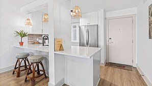 Eine moderne Küche mit weißen Schränken, einem Edelstahl-Kühlschrank und zwei Barhocker mit gemusterten Sitzkissen.