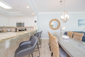 A modern kitchen with white cabinets, a granite countertop, and a built-in microwave. There is a dining area with a glass table and wicker chairs. - Three Bedroom ~ 3 BATH!! ~ Phoenix Gulf Shores *BeachFront* (Gulf Shores)