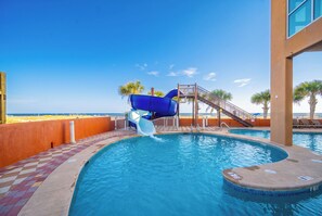 A swimming pool with a water slide, a beach view, and a staircase leading to a deck area.