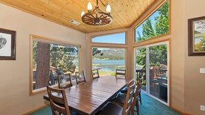 Un comedor con una mesa y sillas de madera, ventanas amplias con vistas a un lago y árboles, y una lámpara de araña.