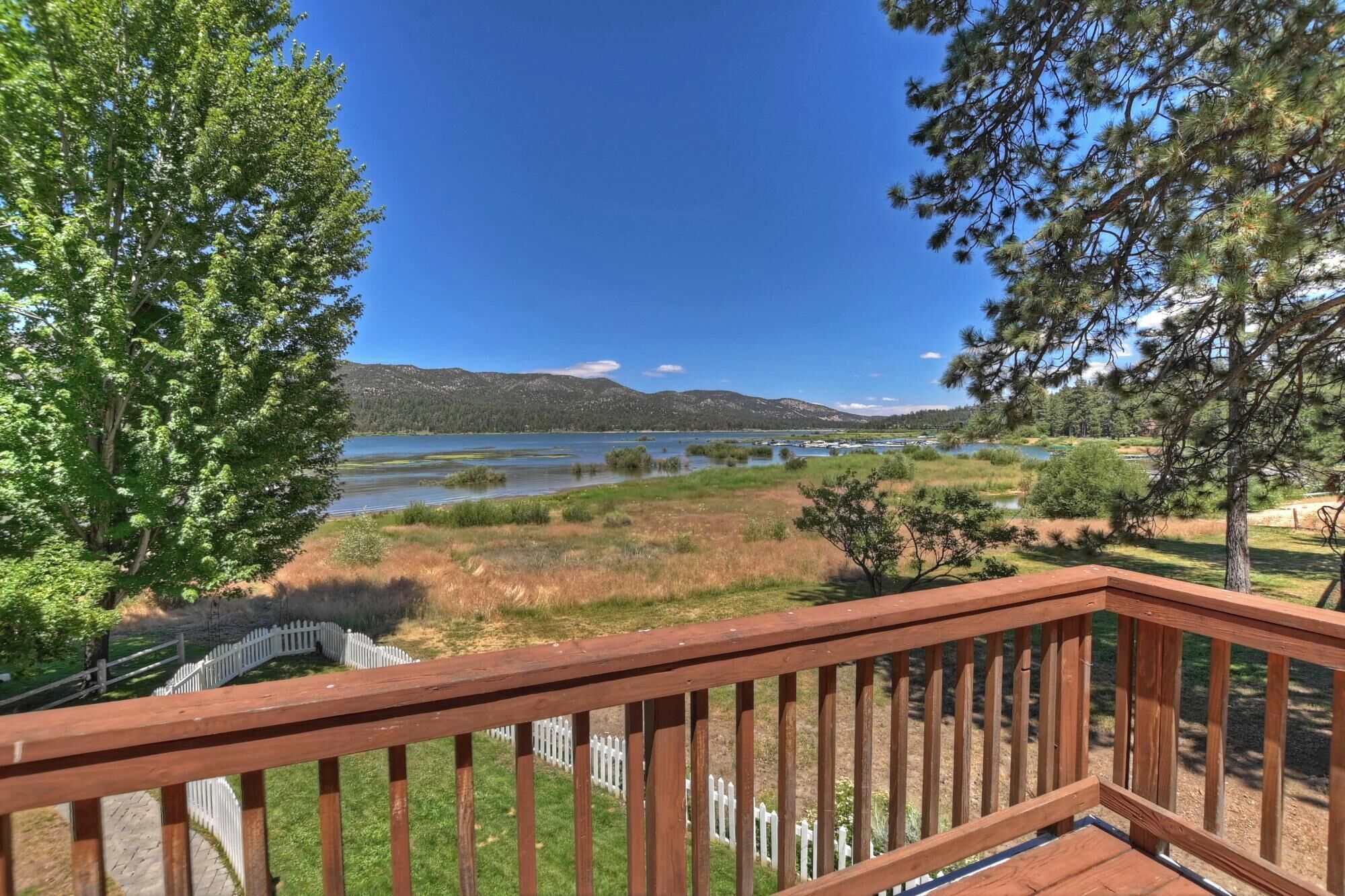 A deck with a view of a lake, surrounded by trees and a white picket fence.