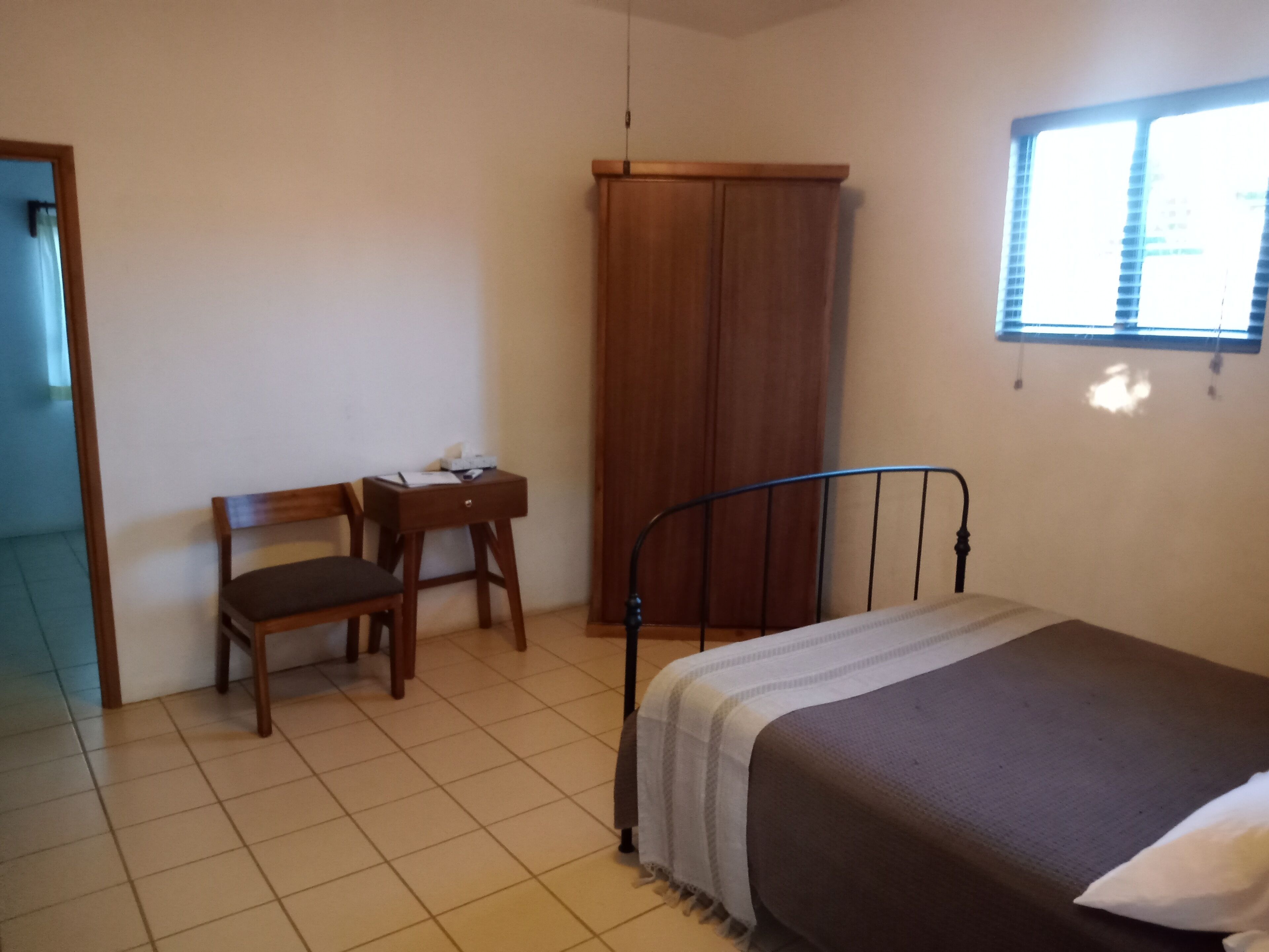 A bedroom with a bed, a wooden desk with a chair, a wardrobe, and a window with blinds.