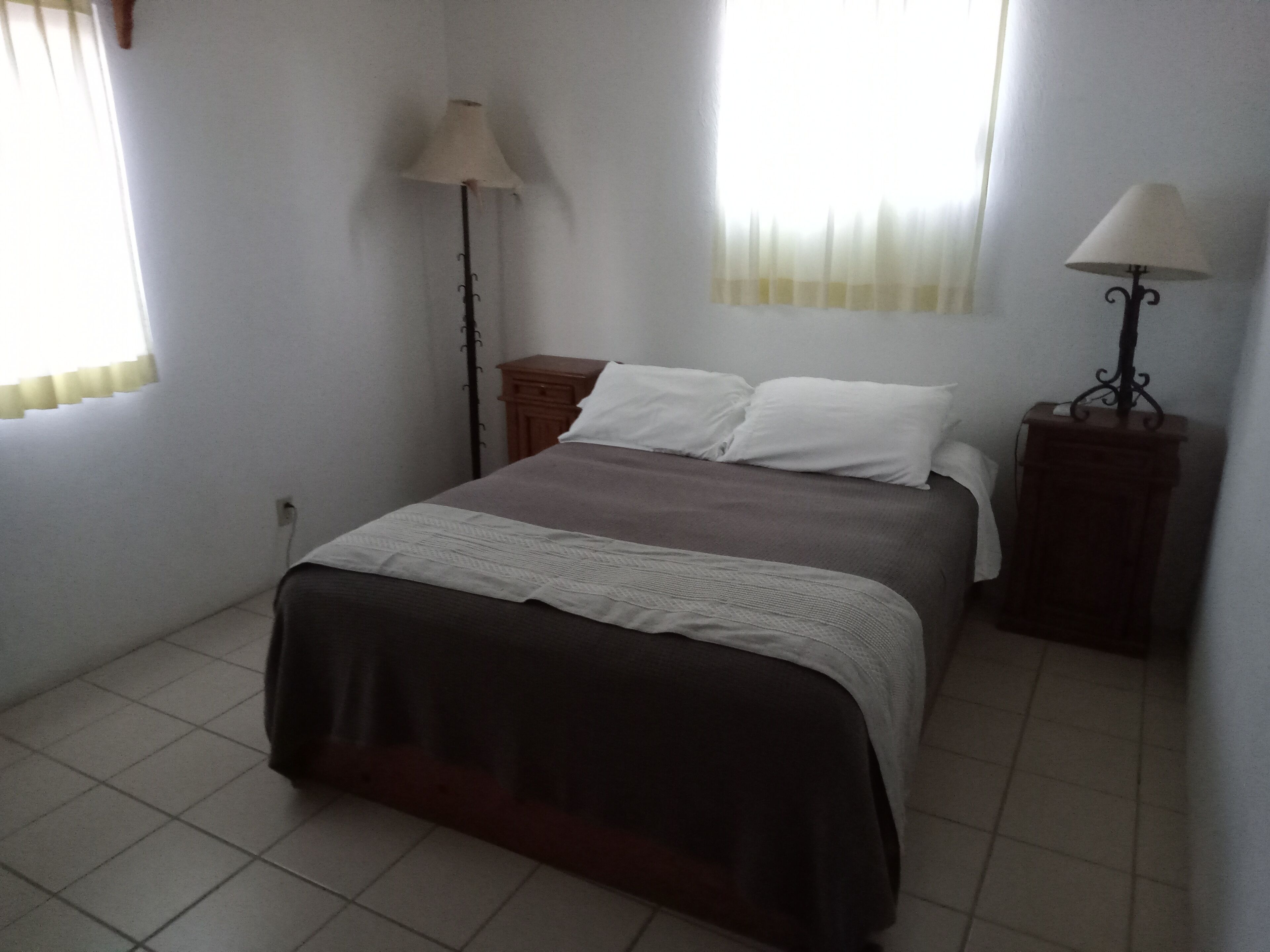 Un dormitorio con cama, dos mesitas de noche, una lámpara de pie y una ventana con cortinas.